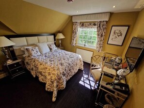Un dormitorio con cama, mesitas de noche, una ventana con cortinas, un radiador, una silla, un escritorio con espejo y un teléfono.