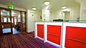 A reception area with a red and white counter, a carpeted floor, and a door marked with an emergency exit sign. - Ayre Hotel (Kirkwall)