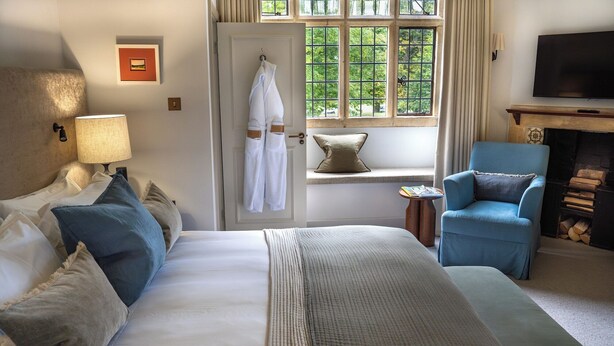 A hotel room with a large bed, a chair, a fireplace, and a window with a view of greenery. - Charingworth Manor (Chipping Campden)