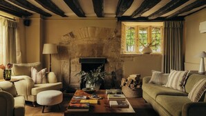 A cozy living room with a fireplace, wooden beams, a sofa, a coffee table, and a window with curtains. - Charingworth Manor (Chipping Campden)