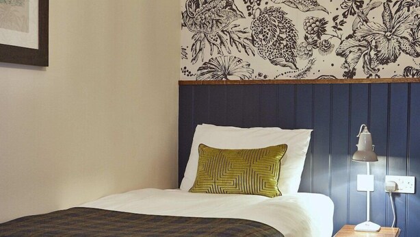Un lit bien fait, recouvert d’une couvre-lit à motifs, agrémenté d’un coussin décoratif et d’une table de chevet avec une lampe.