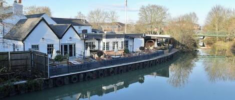 A riverside building with a dock extending into the water, featuring a covered seating area and a small bridge.