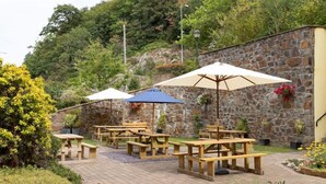 An outdoor patio with wooden picnic tables and umbrellas, surrounded by stone walls and greenery. - Celtic Royal (Caernarfon)