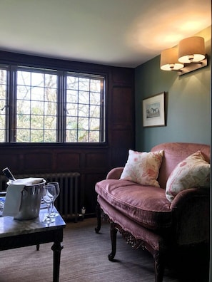 Una habitación con ventana, un radiador, una mesa con jarrón y vasos, una silla acolchada y una lámpara fijada a la pared.
