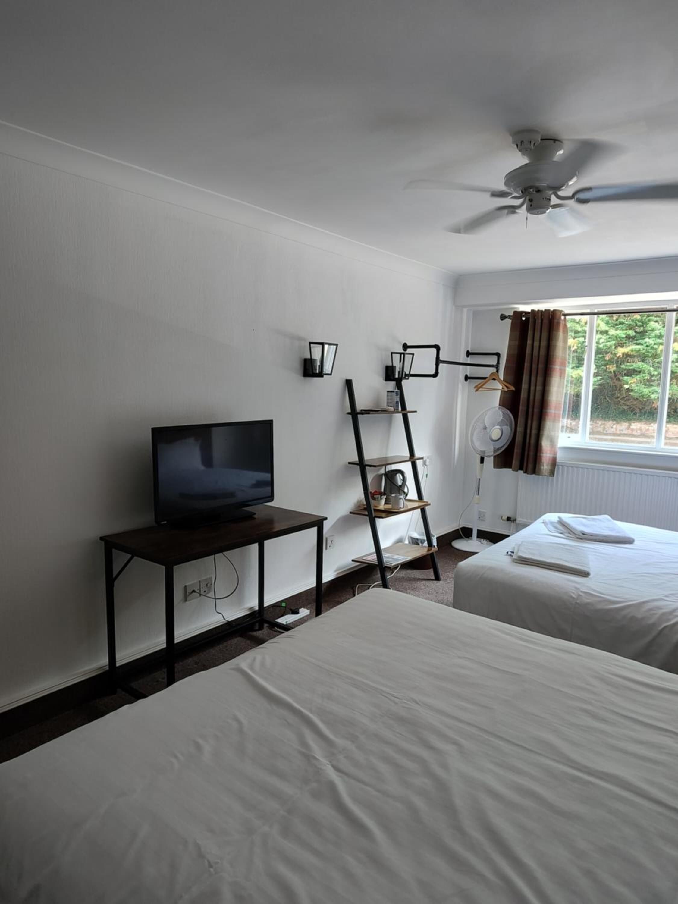 A hotel room with a bed, a TV on a stand, a ceiling fan, and a window with curtains.