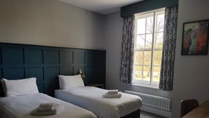 A hotel room with two beds, a window with curtains, a radiator, and a painting on the wall. - Northwick Hotel (Evesham)
