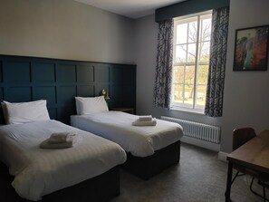 A hotel room with two beds, a window with curtains, a radiator, and a painting on the wall. - Northwick Hotel (Evesham)