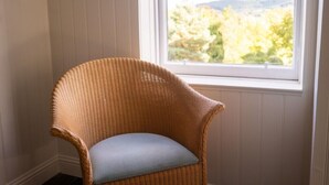 Une chaise en osier avec un coussin clair, dans une pièce aux murs recouverts de lambris blancs et dotée d’une fenêtre donnant sur des arbres.