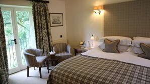 A hotel room with a bed, two chairs, a small table, a nightstand with a lamp, and a window with curtains. - The Bay Tree (Burford)
