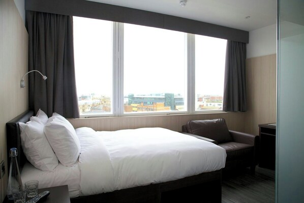 A hotel room with a large bed, a desk, a chair, and a view of the city through large windows. - Z Hotel Liverpool (Liverpool)