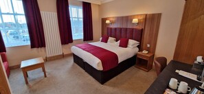 A hotel room with a large bed, a bedside table, a desk with a chair, and a view of the parking lot through the window. - Royal Hotel (Liverpool)
