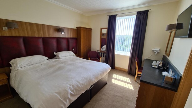 A hotel room with a large bed, a wooden headboard, a desk with a chair, a mirror, and a window with curtains. - Royal Hotel (Liverpool)