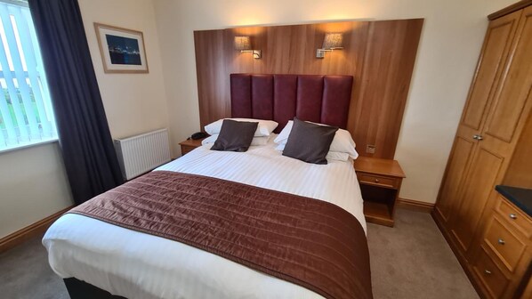 A hotel room with a large bed, a wooden headboard, bedside lamps, a nightstand, a wardrobe, and a window with curtains. - Royal Hotel (Liverpool)