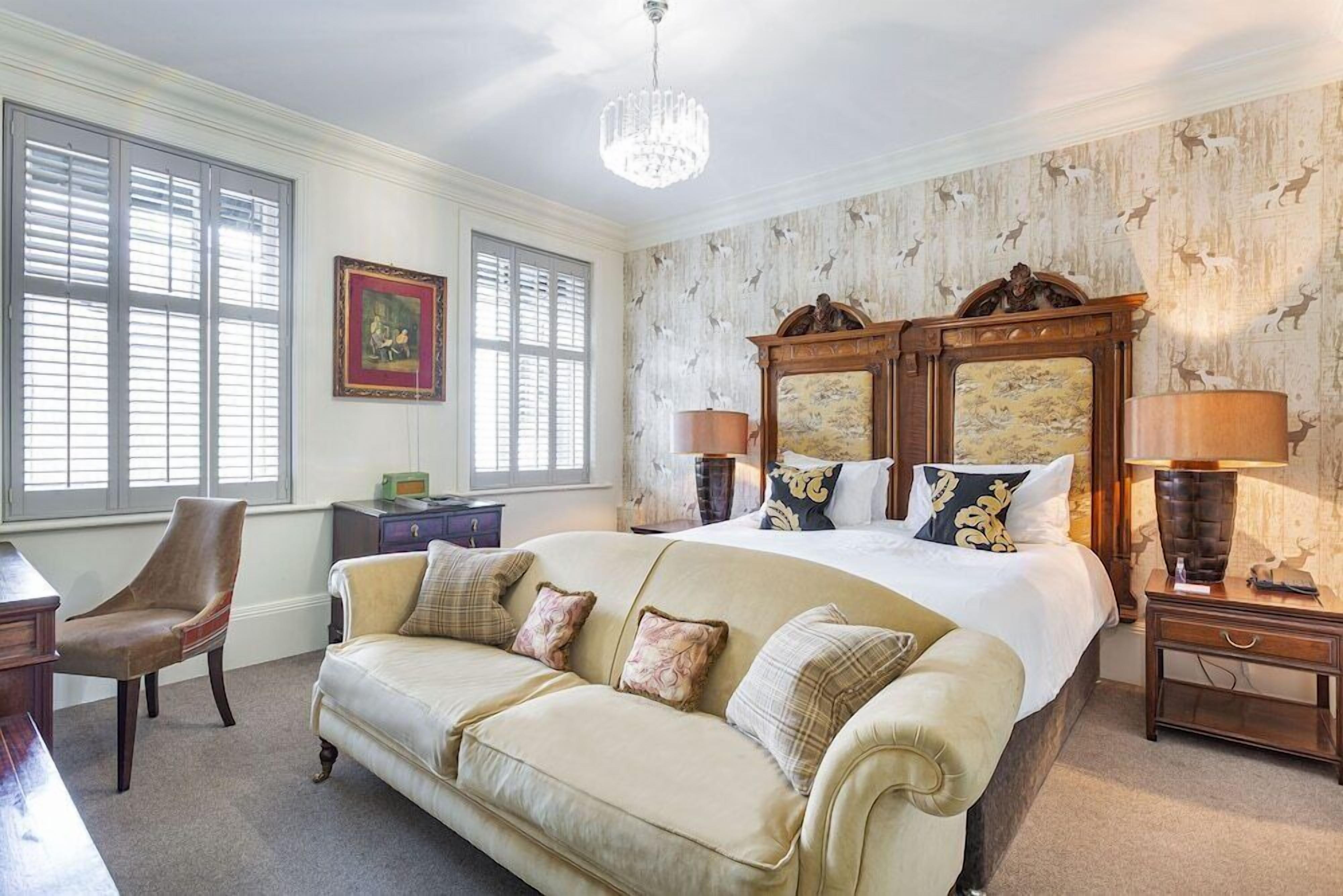A bedroom with a large bed, a sofa, a desk, and a chair. There is a chandelier, a lamp, and a picture frame on the wall.
