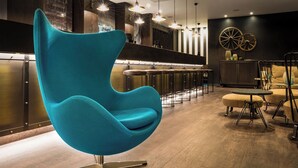 A modern bar area with a teal chair, wooden bar stools, and a large wheel decoration on the wall. - Motel One (Newcastle-upon-Tyne)