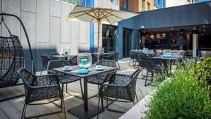 An outdoor patio with wicker chairs and tables, a large umbrella, and a building with blue and brick facades. - Motel One (Newcastle-upon-Tyne)