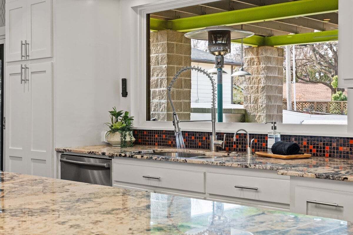 A kitchen with white cabinets, a granite countertop, a window with a green frame, and a stone backsplash.