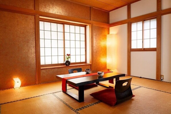 Une pièce de style japonais traditionnel, avec un sol recouvert de tatamis, une table basse surmontée d’une nappe rouge et des portes coulissantes donnant sur un jardin.