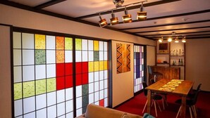 Une pièce dotée d’une grande fenêtre laissant apparaître des motifs colorés, une table à manger en bois avec des chaises, un canapé et un coin bar avec des bouteilles.
