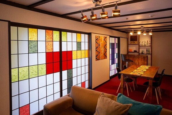 Ein Zimmer mit einem großen Fenster, das bunte Muster zeigt, einem Holzespeisesaal mit Stühlen, einer Couch und einer Bar mit Flaschen.