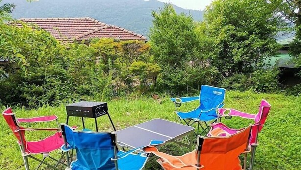 Une table et quatre chaises pliantes colorées, disposées à l’extérieur, dans un décor verdoyant avec une maison en arrière-plan.