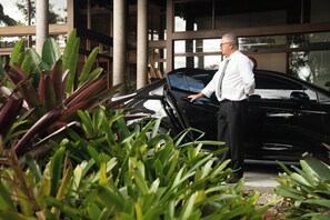 Un hombre de pie junto a un coche negro con la puerta abierta, rodeado de vegetación y un edificio con amplias ventanas.