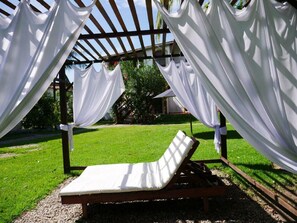 Eine Liege unter einer Pergola mit weißen Vorhängen, umgeben von einer Rasenfläche und Bäumen.