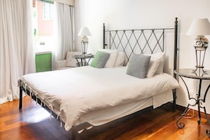 A neatly made bed with a metal headboard, a small side table with a lamp, and a chair with a floral pattern. - Bomtempo II Chales DA Mata EX Villaggio Imperiale Itaipava (Petrópolis)