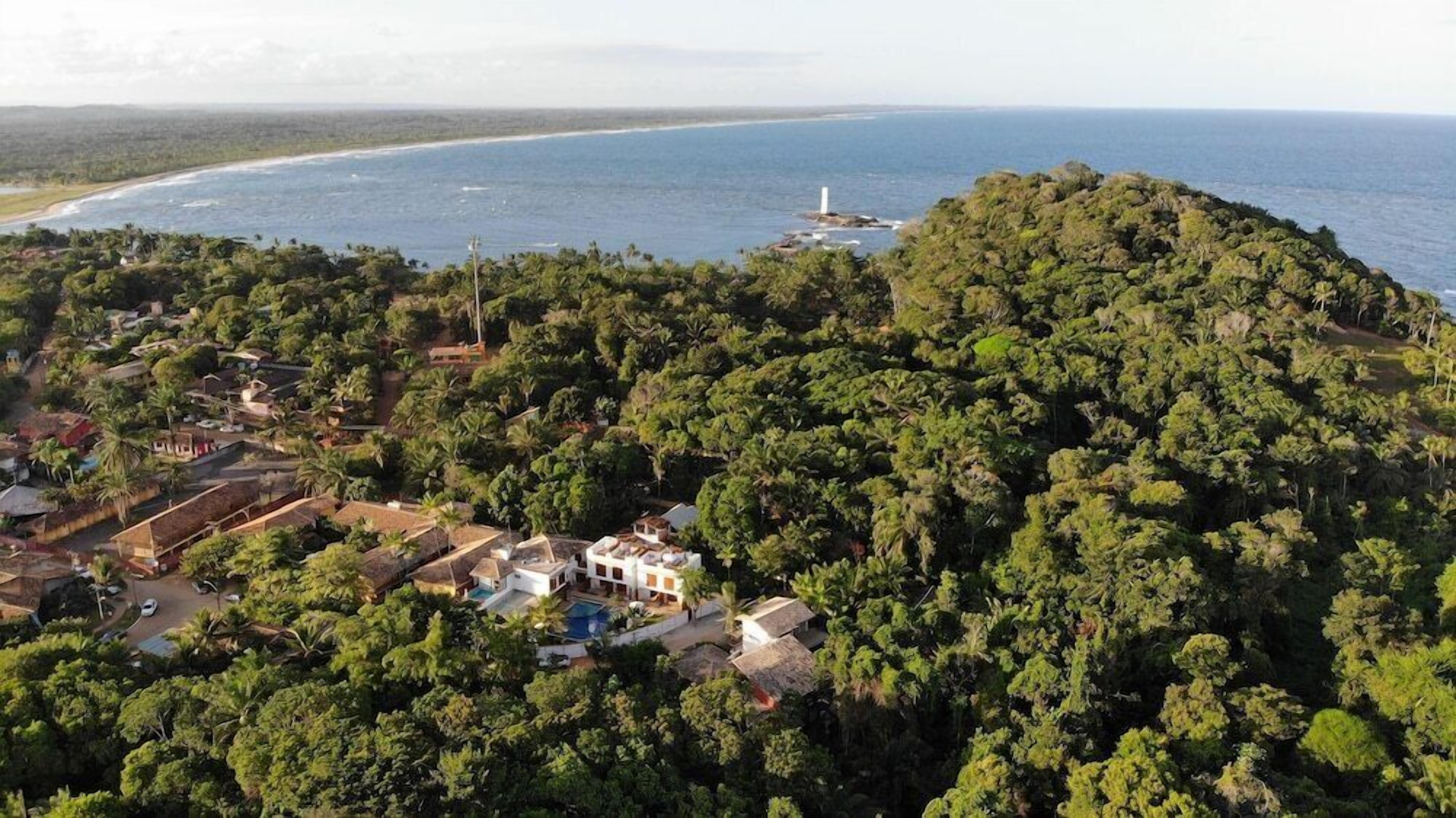 An aerial view of a coastal area with dense greenery, a beach, and a lighthouse.