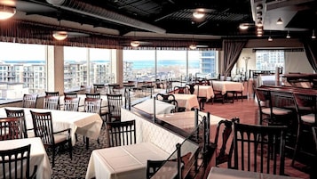 Restaurante com mesas e cadeiras, grandes janelas com vista para a cidade e teto alto com vigas aparentes.