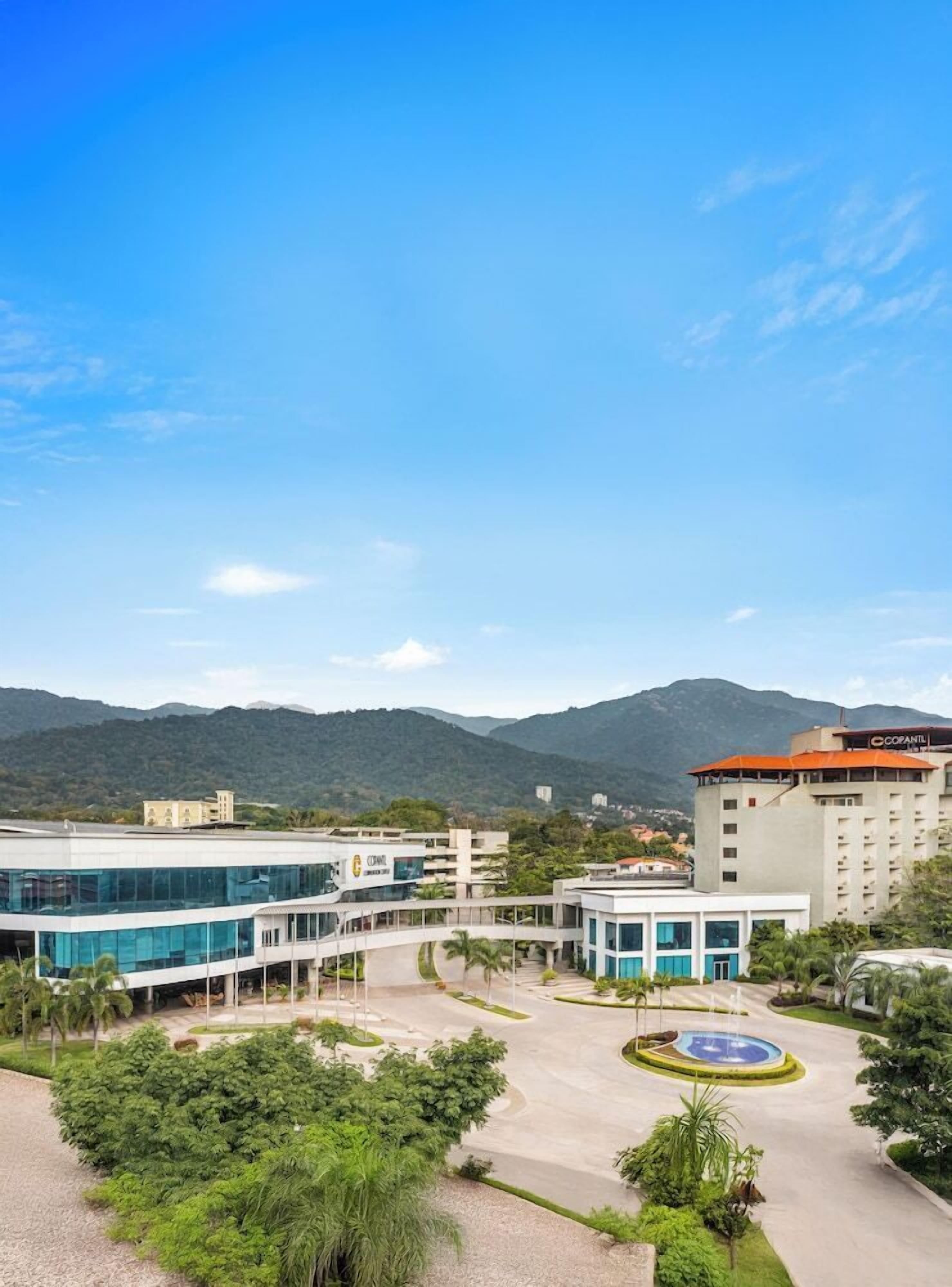 A modern hotel with a central courtyard, surrounded by lush greenery and a mountain backdrop.