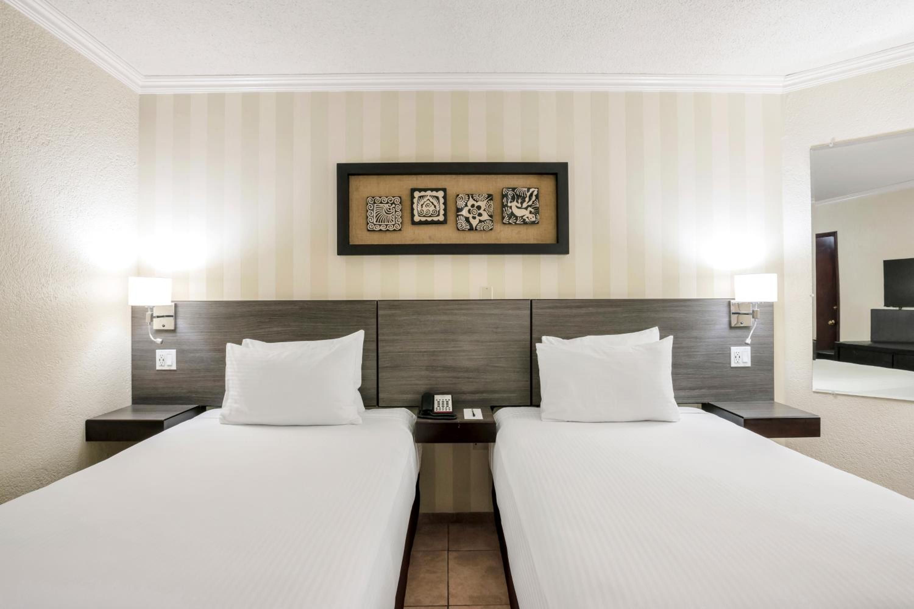 A hotel room with two beds, a headboard, a framed artwork above the beds, and a nightstand with a phone and lamp.
