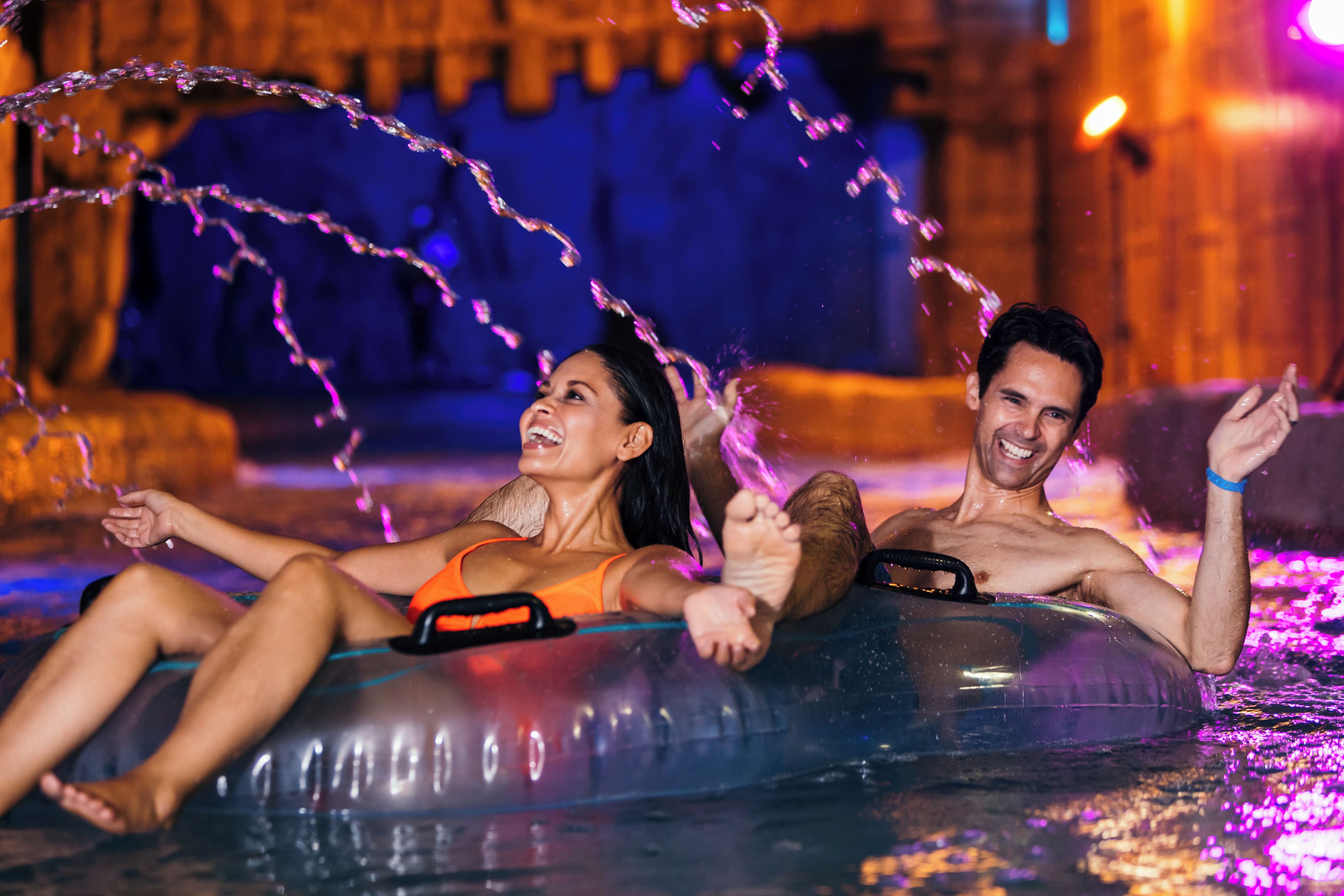 Two people lying on an inflatable tube in a water body, with water jets spraying around them.