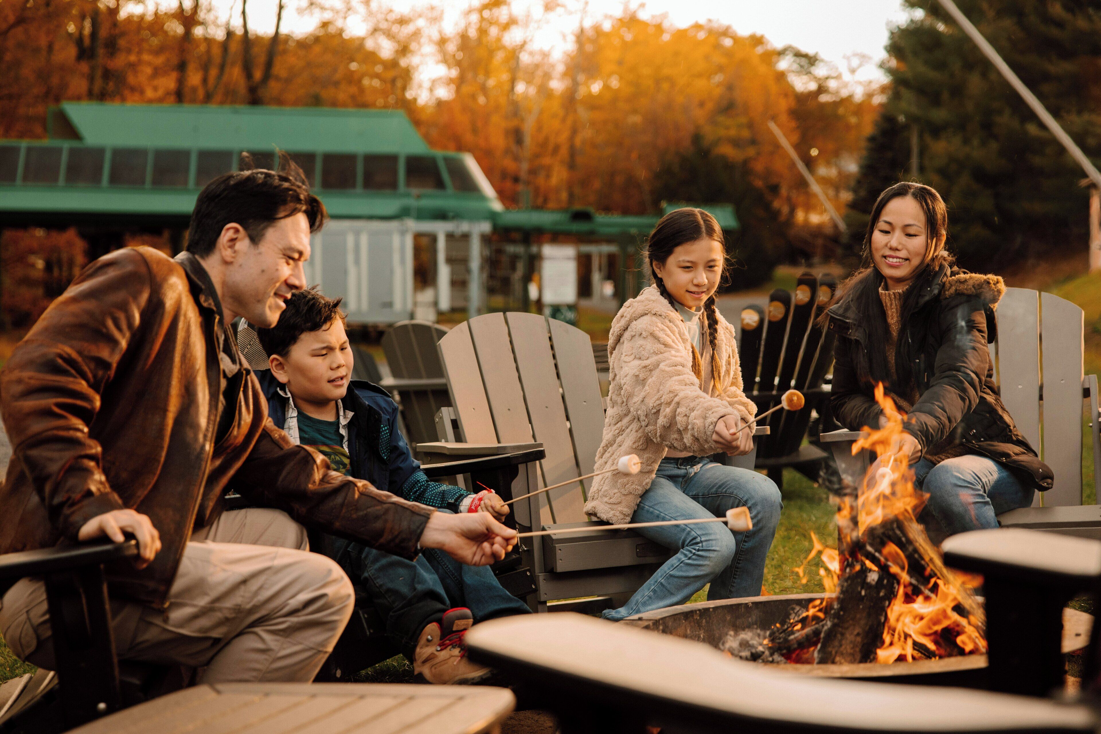 A group of people sitting around a campfire with chairs and tables, in an outdoor setting with trees in the background.