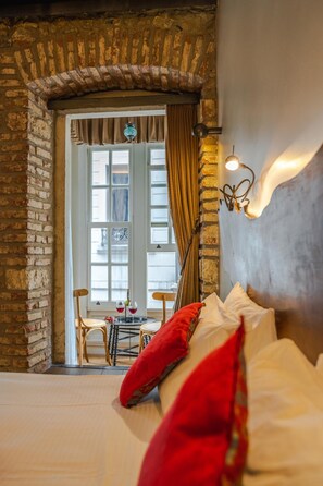 A room with a brick wall, a window with curtains, a bed with red pillows, and a table with a bottle and glasses.