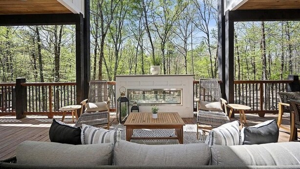 Una terraza al aire libre con mobiliario de mimbre, chimenea y vistas a un bosque.