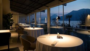 Un restaurante con mesas redondas, amplios ventanales y vistas a montañas y agua.