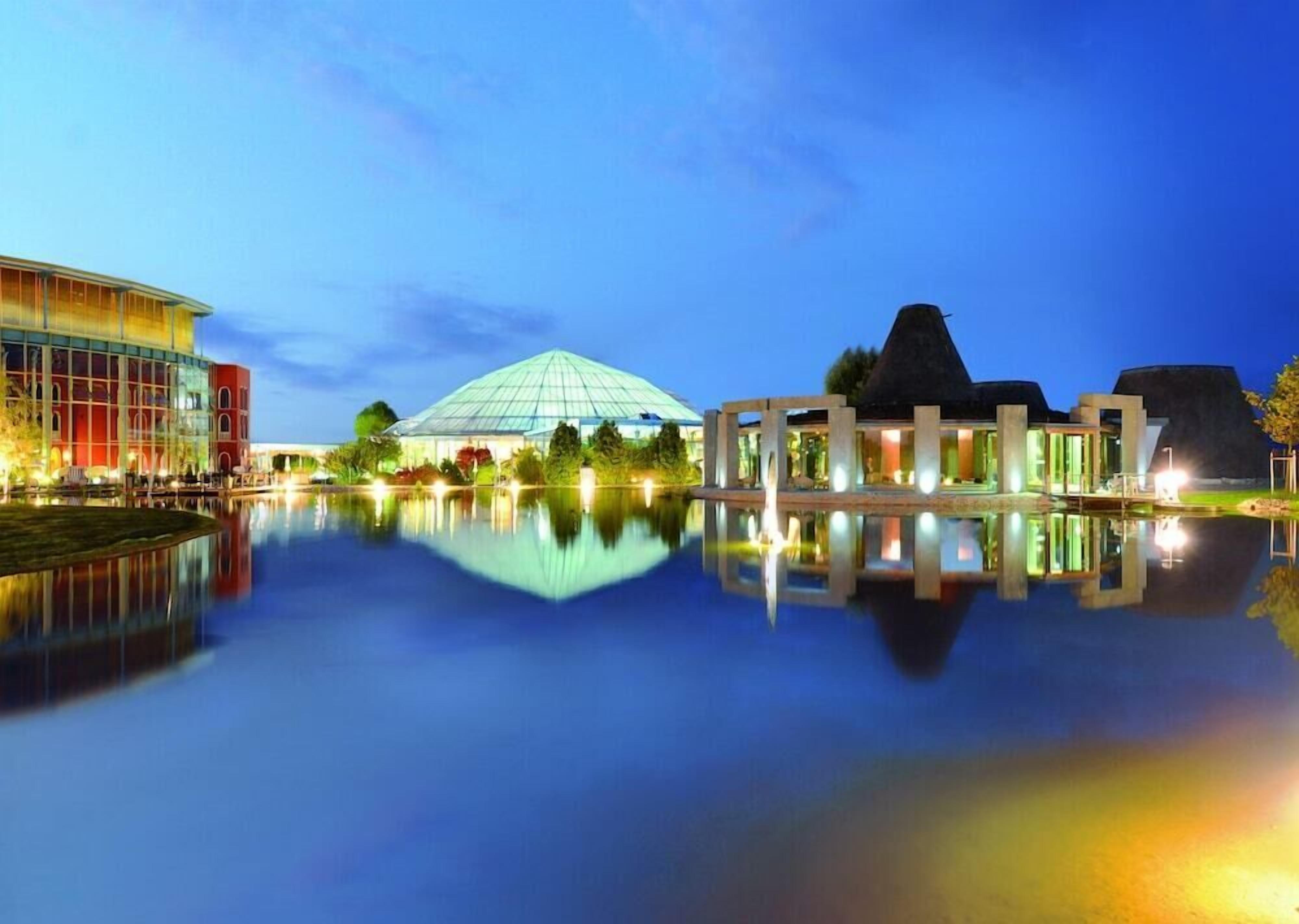 A modern hotel with a glass facade, a large dome structure, and a reflection in the water.