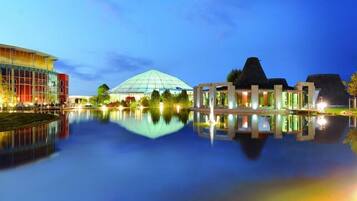 A modern hotel with a glass facade, a large dome structure, and a reflection in the water.