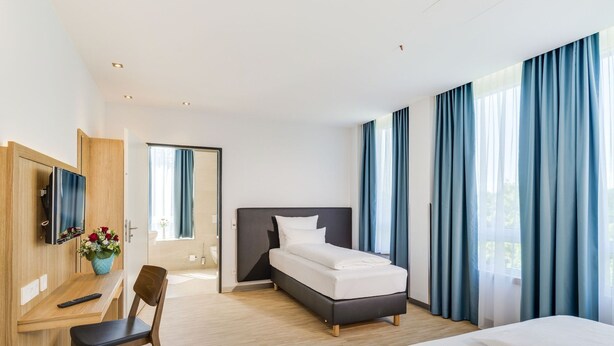 A modern hotel room with a bed, a desk, a chair, a TV, and a bathroom visible through an open door.
