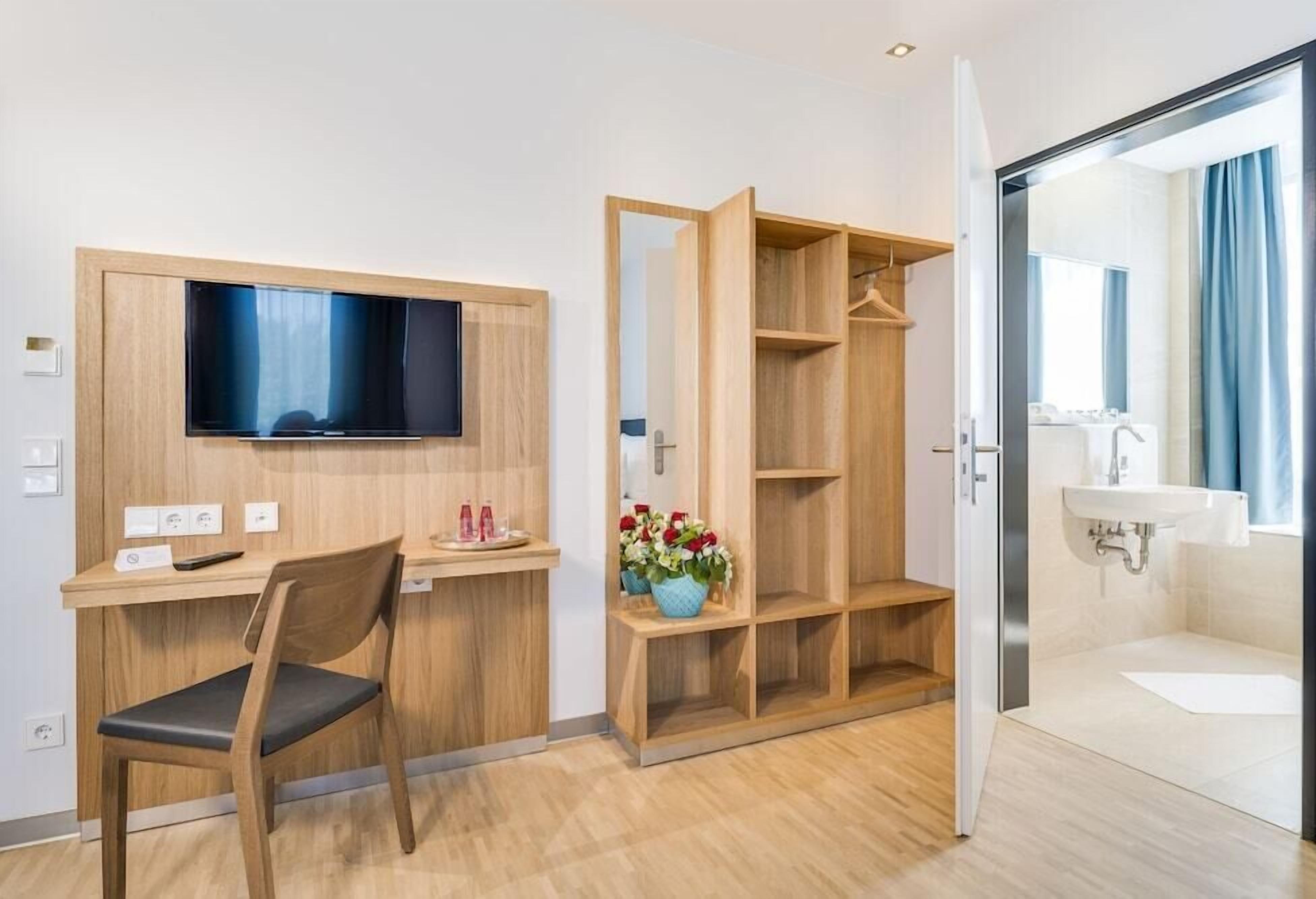 A hotel room with a wooden desk, a chair, a TV mounted on the wall, a wardrobe, a bathroom with a sink and mirror, and a vase with flowers on the desk.
