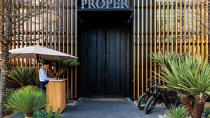 A building entrance with a sign reading 'PROPER,' a wooden counter with a person behind it, and a bicycle parked to the side.