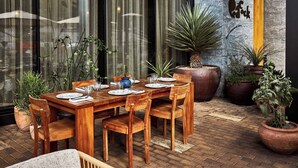 A wooden dining table set with plates, glasses, and cutlery, surrounded by chairs, with potted plants and a large planter in the background.