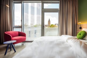 A hotel room with a bed, a red armchair, a small table, and a view of a cityscape including a tower. - Koncept Hotel Josefine (Cologne)