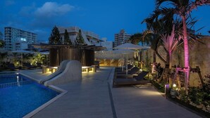 A pool area with a water slide, lounge chairs, and a stone wall with palm trees. - Okinawa Hinode Resort And Hot Spring Hotel (Naha)