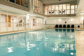 En indendørs swimmingpool med klart vand, en trappeopgang til en balkon og udsigt over interiøret.
