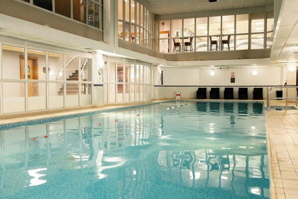 En indendørs swimmingpool med klart vand, en trappeopgang til en balkon og udsigt over interiøret.