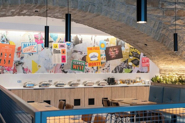 A counter with various posters, a stone wall, and a seating area with tables and chairs. - The Quays Hotel Sheffield (Sheffield)