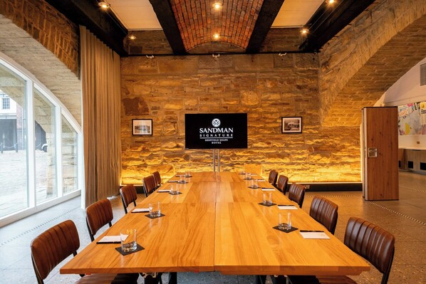 Uma sala de conferências com uma longa mesa de madeira, cadeiras de couro, paredes de pedra e uma grande tela exibindo o nome do hotel.