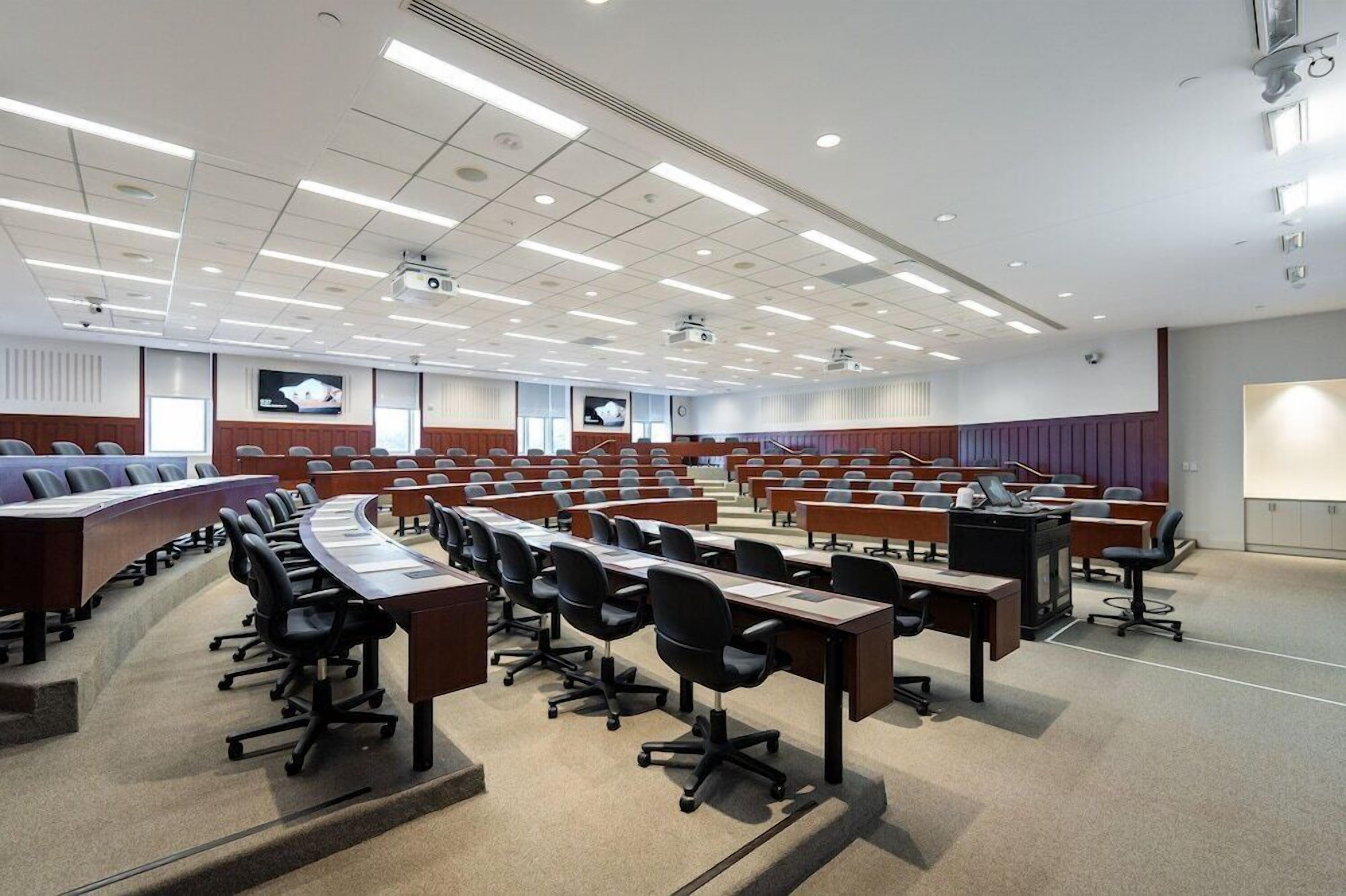 A conference room with multiple rows of tables and chairs, equipped with projectors and screens.